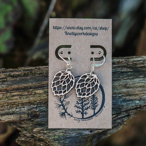 May include: A pair of wooden earrings shaped like pine cones with silver hooks. The earrings are displayed on a brown card with the text "https://www.etsy.com/ca/shop/Knottyworkdesigns" and "Locally handcrafted, Vancouver, BC".