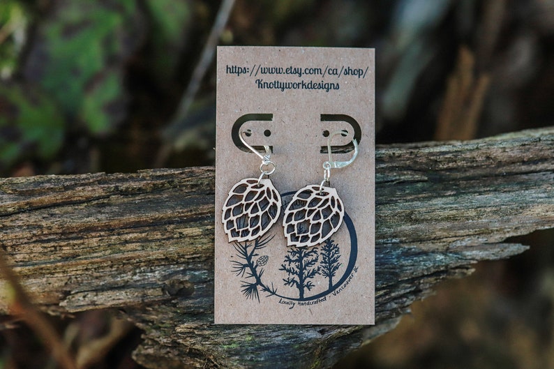 May include: A pair of wooden earrings shaped like pine cones with silver hooks. The earrings are displayed on a brown card with the text "https://www.etsy.com/ca/shop/Knottyworkdesigns" and "Locally handcrafted, Vancouver, BC".