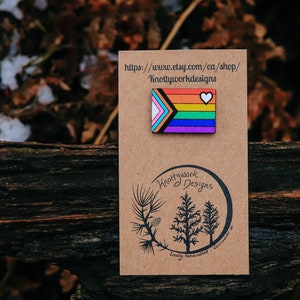May include: A wooden pin with a rainbow flag design and a white heart. The pin is on a brown card with the text "Knottywork Designs" and a website address.