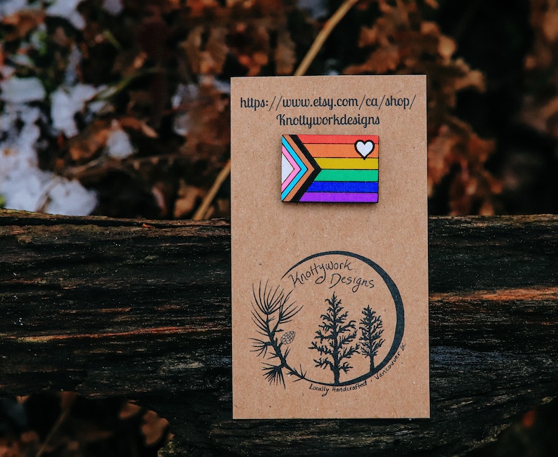 May include: A wooden pin with a rainbow flag design and a white heart. The pin is on a brown card with the text "Knottywork Designs" and a website address.