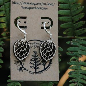 May include: A pair of silver earrings with a wooden leaf design. The leaves are brown and have a detailed pattern. The earrings are on a brown card with the text "Knottyworkdesigns" and a website address.