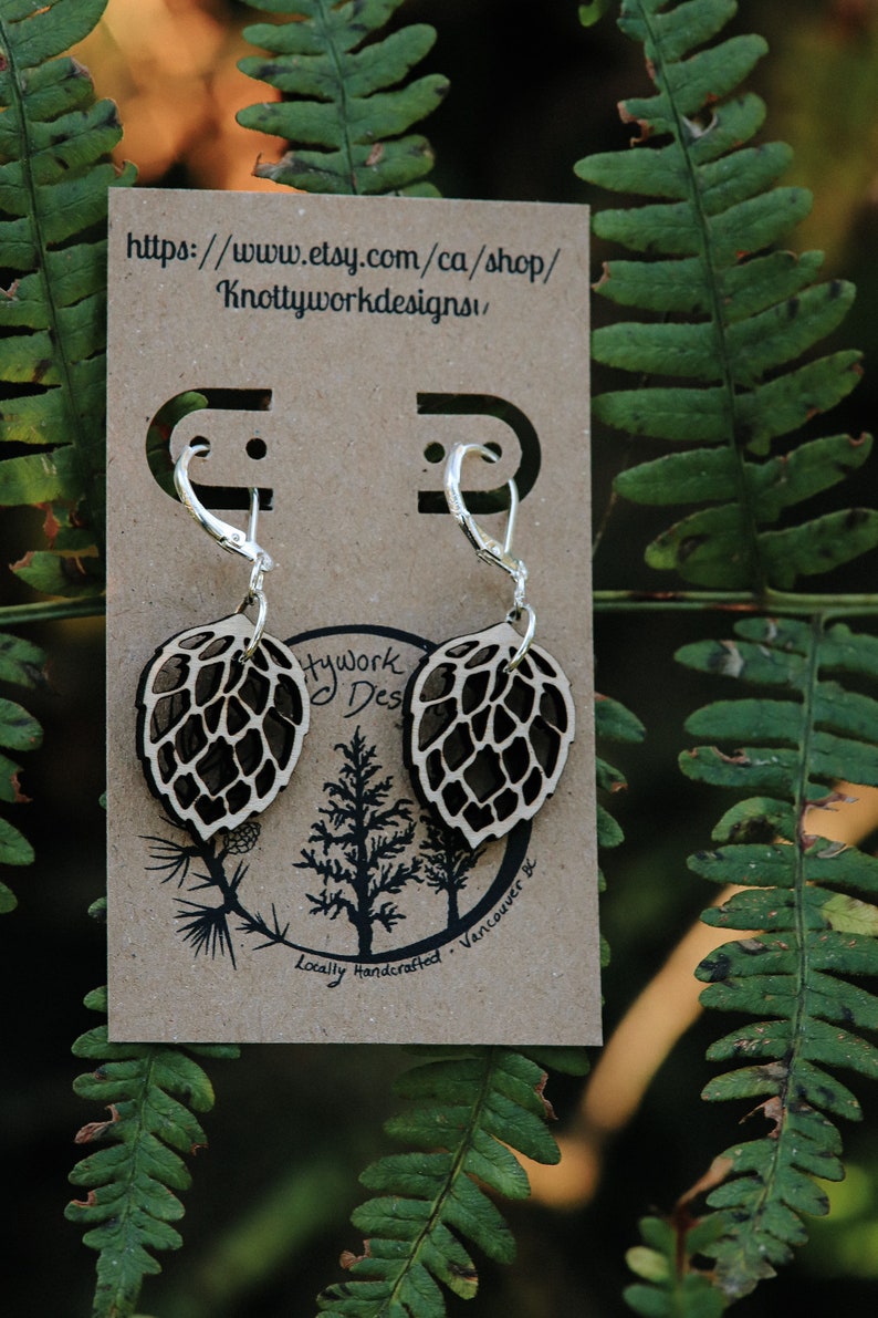 May include: A pair of silver earrings with a wooden leaf design. The leaves are brown and have a detailed pattern. The earrings are on a brown card with the text "Knottyworkdesigns" and a website address.