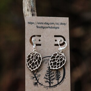 May include: A pair of wooden earrings shaped like pine cones with silver hooks. The earrings are displayed on a brown card with the text "https://www.etsy.com/ca/shop/Knottyworkdesigns" and "Locally handcrafted, Vancouver, BC".