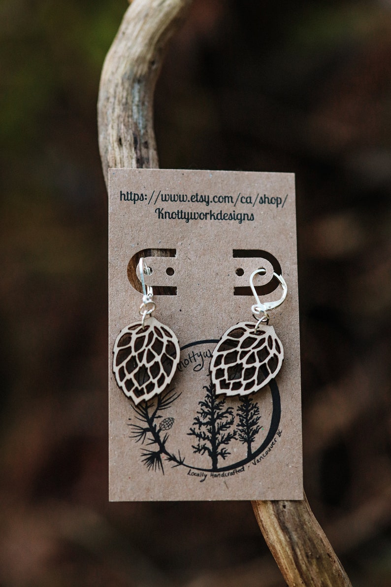 May include: A pair of wooden earrings shaped like pine cones with silver hooks. The earrings are displayed on a brown card with the text "https://www.etsy.com/ca/shop/Knottyworkdesigns" and "Locally handcrafted, Vancouver, BC".