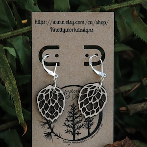 May include: A pair of wooden earrings shaped like pine cones with silver hoop ear wires. The earrings are displayed on a brown card with the text "Knottyworkdesigns" and a website address.