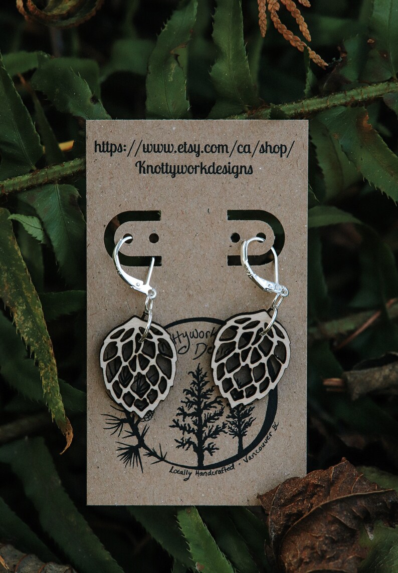 May include: A pair of wooden earrings shaped like pine cones with silver hoop ear wires. The earrings are displayed on a brown card with the text "Knottyworkdesigns" and a website address.