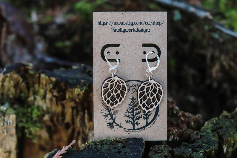 May include: A pair of wooden earrings shaped like a hop cone, with silver hooks. The earrings are displayed on a brown card with the text "https://www.etsy.com/ca/shop/Knottyworkdesigns" and "Lovely handcrafted - Vancouver, BC".