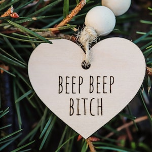Może przedstawiać: Drewniana ozdoba w kształcie serca z napisem "BEEP BEEP BITCH" wisząca na gałęzi sosny.