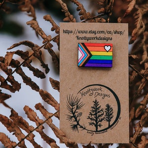 May include: A wooden pin featuring a rainbow pride flag with a white heart in the top right corner. The pin is on a brown card with the text "Knottywork Designs" and a website address.