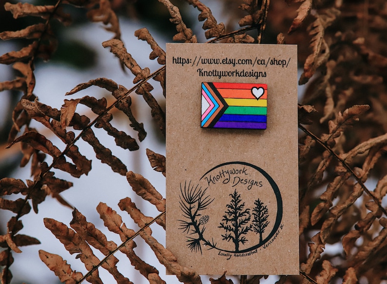 May include: A wooden pin featuring a rainbow pride flag with a white heart in the top right corner. The pin is on a brown card with the text "Knottywork Designs" and a website address.