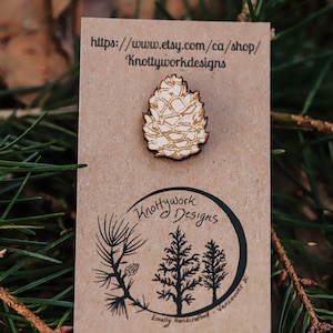 May include: A wooden pin shaped like a pine cone. The pin is on a brown card with the text "Knottywork Designs" and a website address.