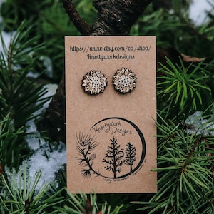 May include: A pair of wooden sunflower earrings on a brown card with the text "Knottywork Designs" and a website address. The earrings are a light brown color with a white design.