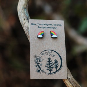 Pu&ograve; includere: Un paio di orecchini arcobaleno e nuvola su una carta marrone con il testo "Knottywork Designs" e un indirizzo di sito web.