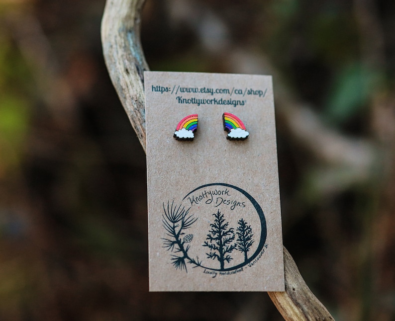 Pu&ograve; includere: Un paio di orecchini arcobaleno e nuvola su una carta marrone con il testo "Knottywork Designs" e un indirizzo di sito web.