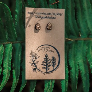 K&ouml;nnte beinhalten: Ein Paar Holz-Kiefernzapfen-Ohrringe auf einer braunen Karte mit dem Text "Knottywork Designs" und der Website-Adresse "https://www.etsy.com/ca/shop/Knottyworkdesigns".