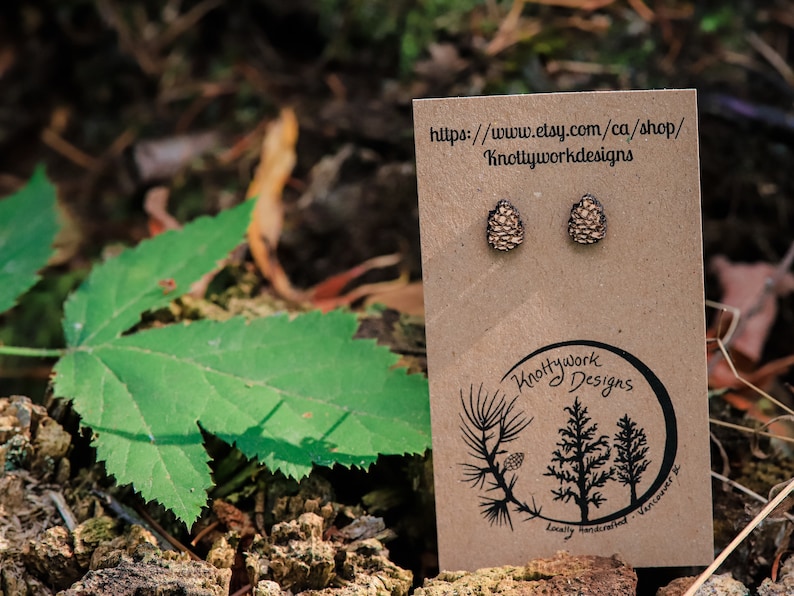 K&ouml;nnte beinhalten: Ein Paar braune und beigefarbene Holz-Kiefernzapfen-Ohrstecker auf einer braunen Karte mit dem Text "Knottywork Designs" und einer Website-Adresse.