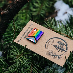May include: A rainbow-colored wooden pin with a white heart on top. The pin is on a brown card with the text "Knottywork Designs" and a website address.