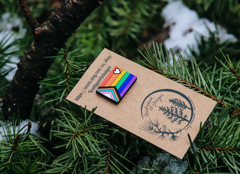 May include: A rainbow-colored wooden pin with a white heart on top. The pin is on a brown card with the text "Knottywork Designs" and a website address.
