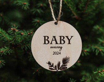 Op de afbeelding: Een ronde houten ornament met de tekst "BABY arriving 2024" en een dennentak ontwerp.