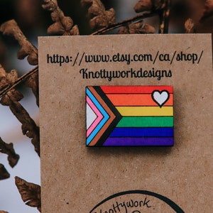 May include: A wooden pin with a rainbow flag design and a white heart. The pin is on a brown card with the text "https://www.etsy.com/ca/shop/Knottyworkdesigns".
