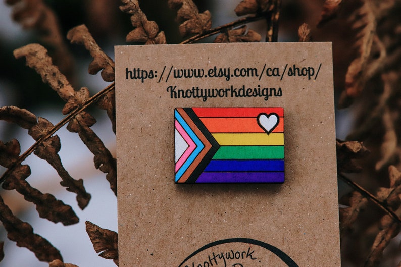 May include: A wooden pin with a rainbow flag design and a white heart. The pin is on a brown card with the text "https://www.etsy.com/ca/shop/Knottyworkdesigns".