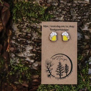 May include: A pair of wooden beer mug earrings with a yellow mug and white foam. The earrings are on a brown card with the text "Knottywork Designs" and a website address.