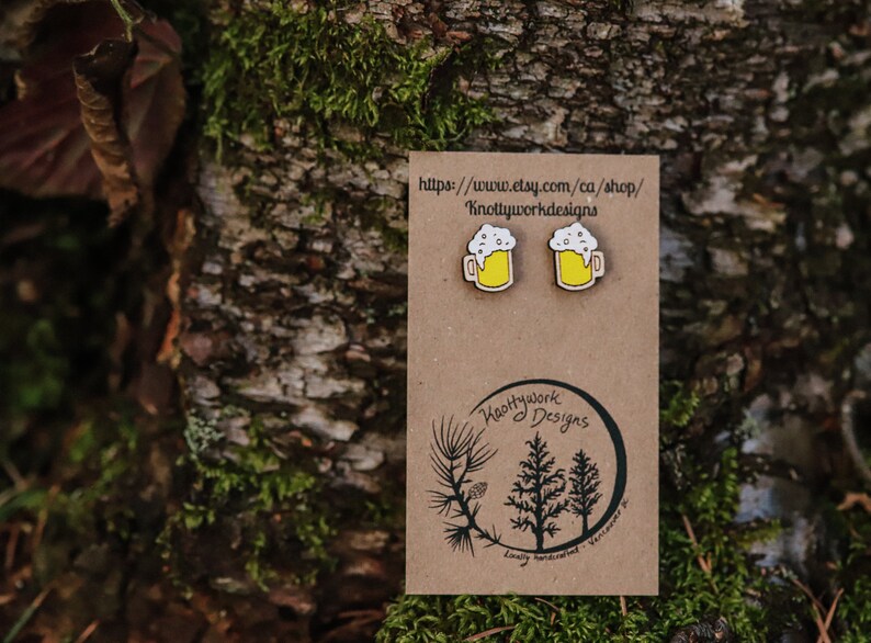 May include: A pair of wooden beer mug earrings with a yellow mug and white foam. The earrings are on a brown card with the text "Knottywork Designs" and a website address.