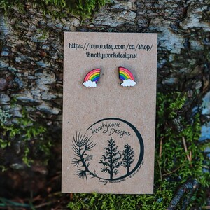Pu&ograve; includere: Un paio di orecchini in legno a forma di arcobaleno e nuvola su una carta marrone con il testo "Knottywork Designs" e un indirizzo di sito web.