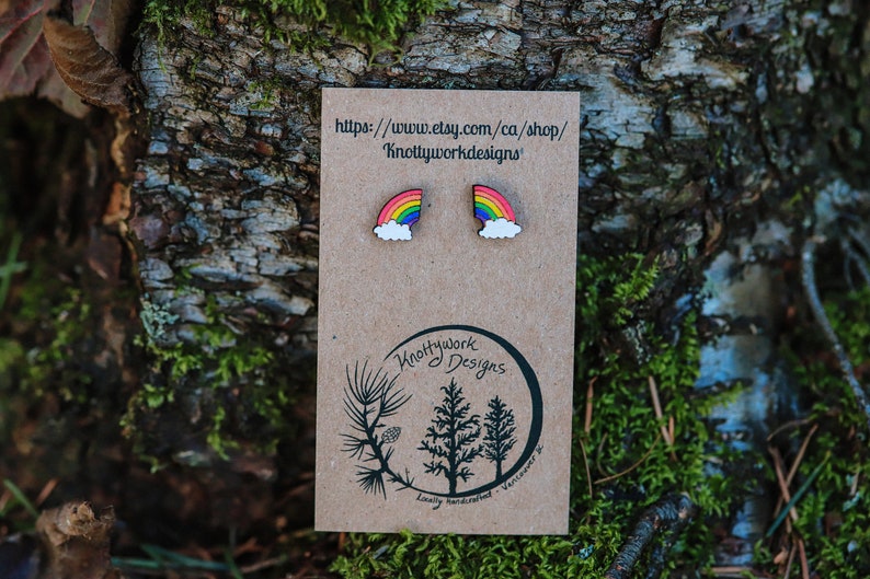 Pu&ograve; includere: Un paio di orecchini in legno a forma di arcobaleno e nuvola su una carta marrone con il testo "Knottywork Designs" e un indirizzo di sito web.
