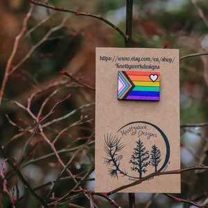 May include: A wooden pin with a rainbow flag design and a white heart. The pin is on a brown card with the text "Knottywork Designs" and a website address.