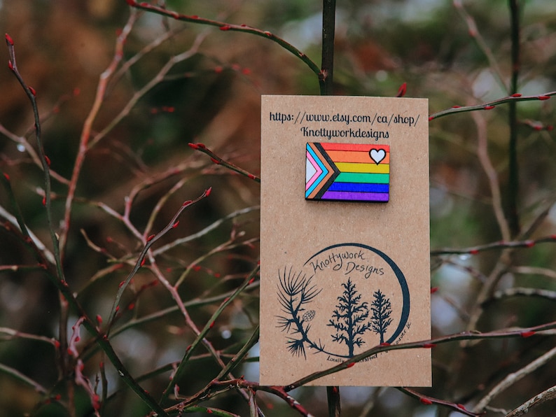 May include: A wooden pin with a rainbow flag design and a white heart. The pin is on a brown card with the text "Knottywork Designs" and a website address.