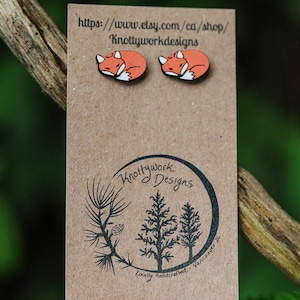 Könnte beinhalten: Ein Paar Holzohrringe in Form eines Fuchses mit orangefarbenem Fell und weißen Akzenten. Die Ohrringe werden auf einer braunen Karte mit dem Text "Knottywork Designs" und "Lokale Handarbeit - Vancouver BC" präsentiert.