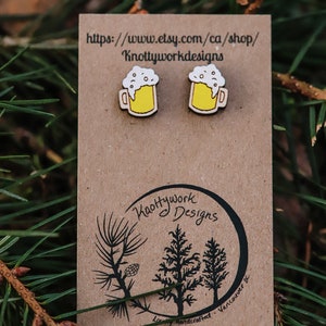 May include: A pair of wooden earrings shaped like beer mugs with yellow liquid and white foam. The earrings are on a brown card with the text "Knottywork Designs" and a website address.