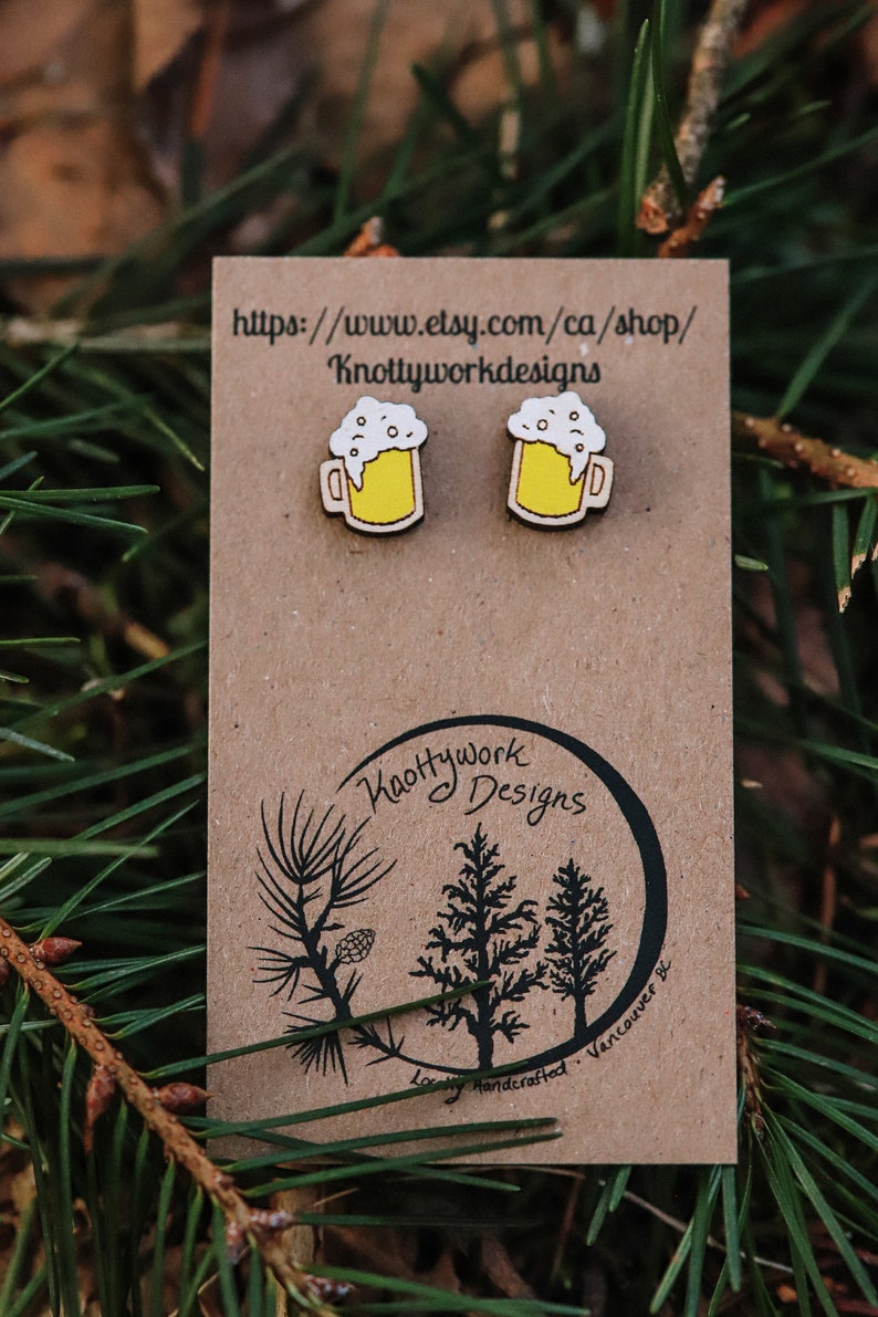 May include: A pair of wooden earrings shaped like beer mugs with yellow liquid and white foam. The earrings are on a brown card with the text "Knottywork Designs" and a website address.