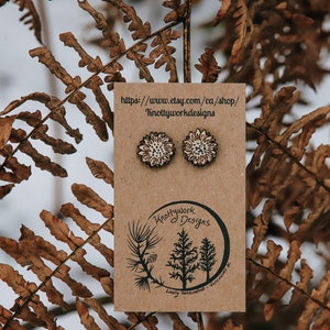 May include: A pair of wooden sunflower earrings on a brown card with the text "Knottywork Designs" and a website address. The earrings are white with a brown center.