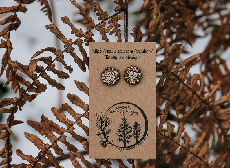 May include: A pair of wooden sunflower earrings on a brown card with the text "Knottywork Designs" and a website address. The earrings are white with a brown center.