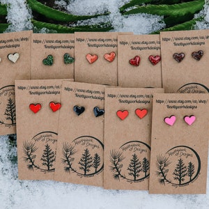 May include: An assortment of heart-shaped earrings displayed on brown cards with the text "Knottywork Designs" and a website address. The earrings are made of wood and have glitter embedded in the paint. The colours of the earrings include red, pink, black, green, and white.