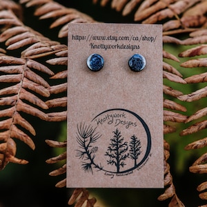 May include: A pair of small, round, black and blue earrings with a speckled finish. The earrings are displayed on a brown card with the text "Knottywork Designs" and a website address.