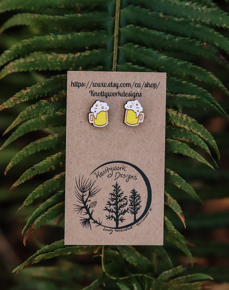 May include: A pair of wooden beer mug earrings with yellow and white paint. The earrings are displayed on a brown card with the text "Knottywork Designs" and a website address.