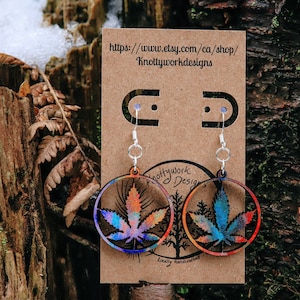 Peut inclure: Boucles d'oreilles rondes avec un motif de feuille de cannabis multicolore, avec des nuances de bleu, d'orange et de violet. Les boucles d'oreilles sont suspendues à des crochets argentés et présentées sur une carte marron.