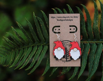 May include: A pair of red and white wooden gnome earrings with silver hooks. The earrings are displayed on a brown card with the text "Knottywork Designs" and a website address.