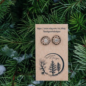 May include: A pair of wooden sunflower earrings with a white and brown design. The earrings are displayed on a brown card with the text "Knottywork Designs" and a website address.