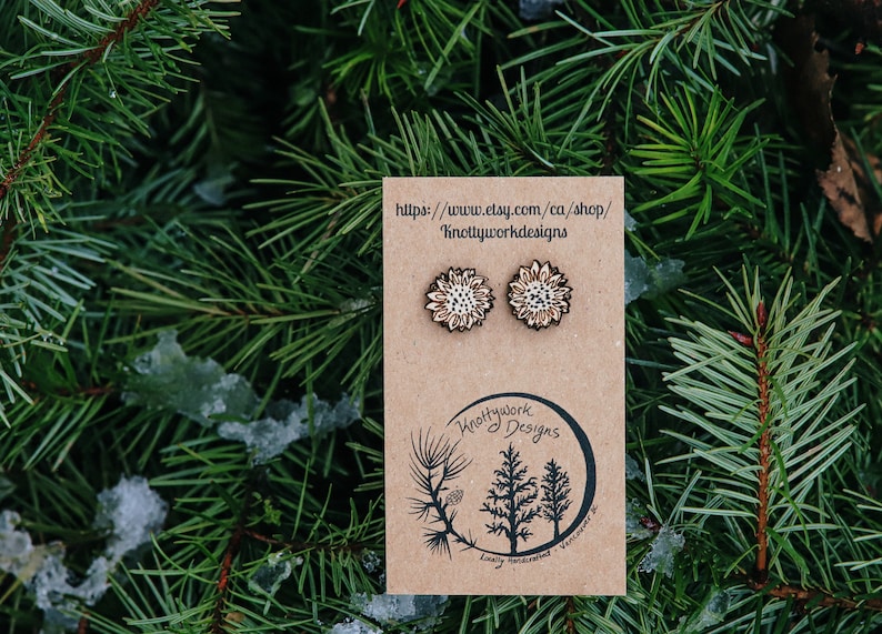May include: A pair of wooden sunflower earrings with a white and brown design. The earrings are displayed on a brown card with the text "Knottywork Designs" and a website address.