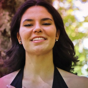 Pu&ograve; includere: Una donna con i capelli scuri e un sorriso caldo, indossa un top nero con spalline, &egrave; in piedi davanti a uno sfondo sfocato di alberi.
