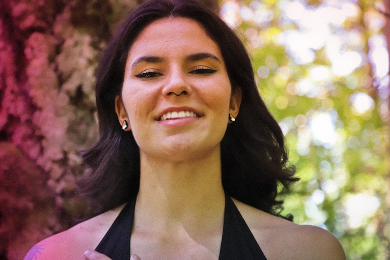 Pu&ograve; includere: Una donna con i capelli scuri e un sorriso caldo, indossa un top nero con spalline, &egrave; in piedi davanti a uno sfondo sfocato di alberi.