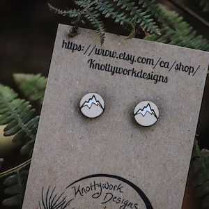 May include: A pair of wooden earrings featuring a mountain design. The earrings are displayed on a brown card with the text "https://www.etsy.com/ca/shop/Knottyworkdesigns".