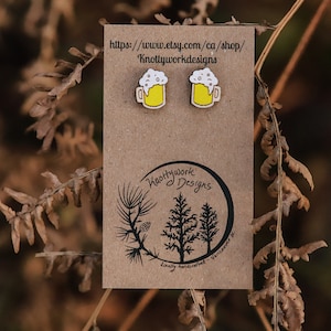 May include: A pair of wooden earrings shaped like beer mugs with white foam and yellow liquid. The earrings are on a brown card with the text "Knottywork Designs" and a website address.