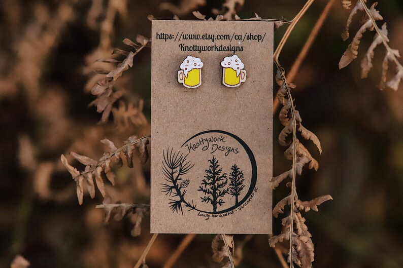 May include: A pair of wooden earrings shaped like beer mugs with white foam and yellow liquid. The earrings are on a brown card with the text "Knottywork Designs" and a website address.