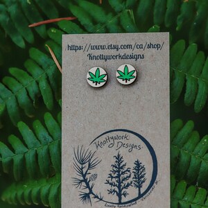 May include: A pair of wooden earrings with green marijuana leaf designs. The earrings are on a brown card with the text "Knottywork Designs" and a website address.