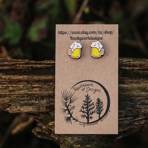 May include: A pair of wooden beer mug earrings with a white foam top and yellow liquid. The earrings are on a brown card with the text "Knottywork Designs" and a website address.